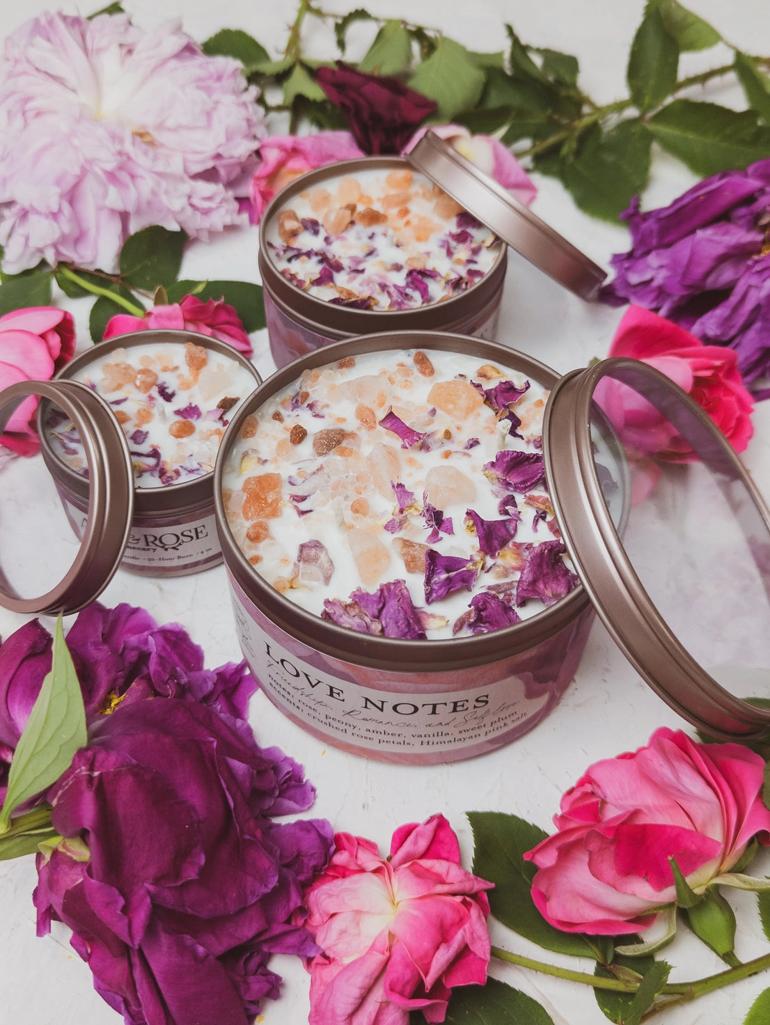 Candle tins labeled 'Love Notes' surrounded by pink and purple flowers on a white background