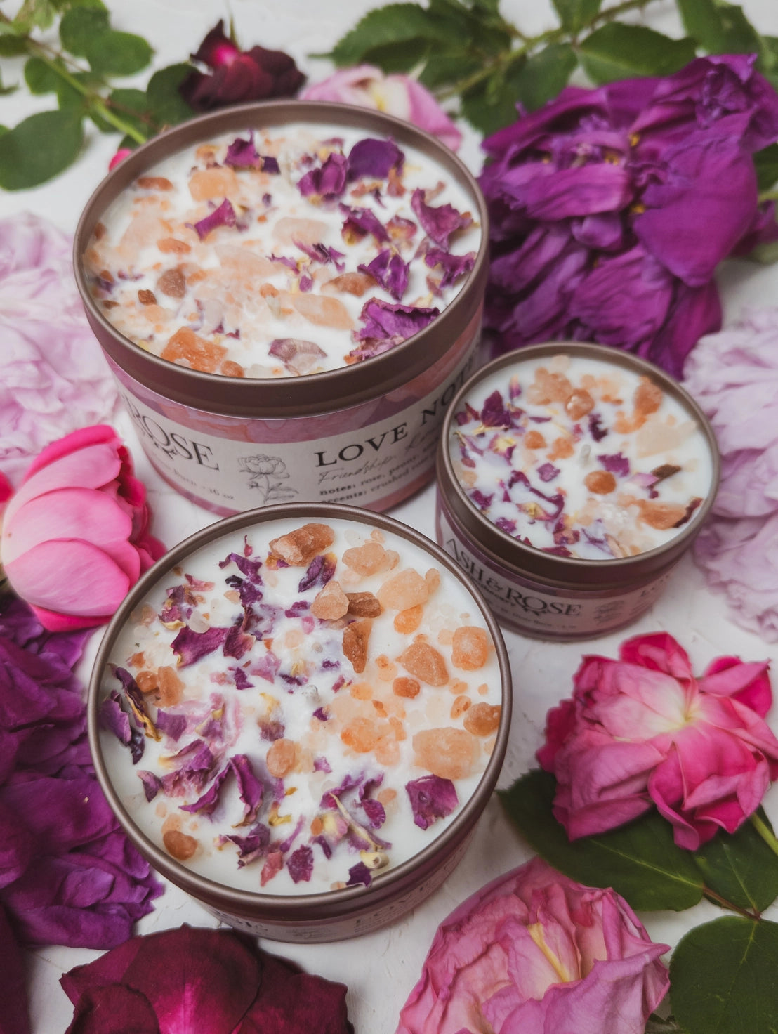 Three tins of 'Love Muse' body scrub on a bed of pink and purple flowers.