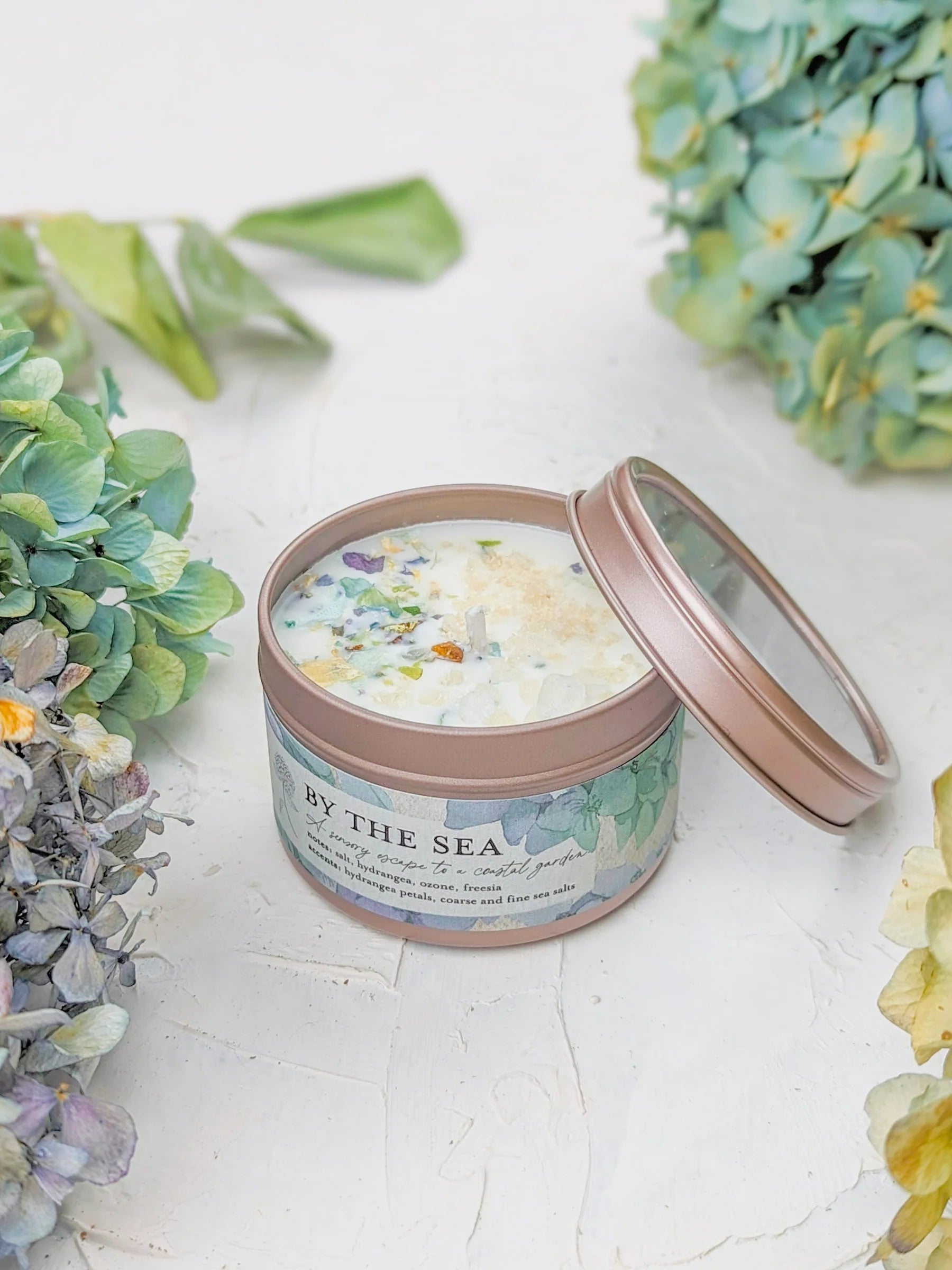 Candle in a tin labeled 'By the Sea' surrounded by flowers on a light surface