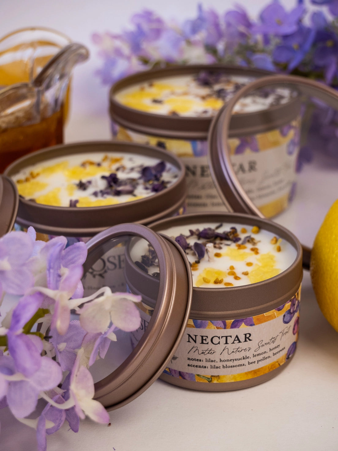 Candles in tins with 'Nectar' label surrounded by purple flowers and a lemon.