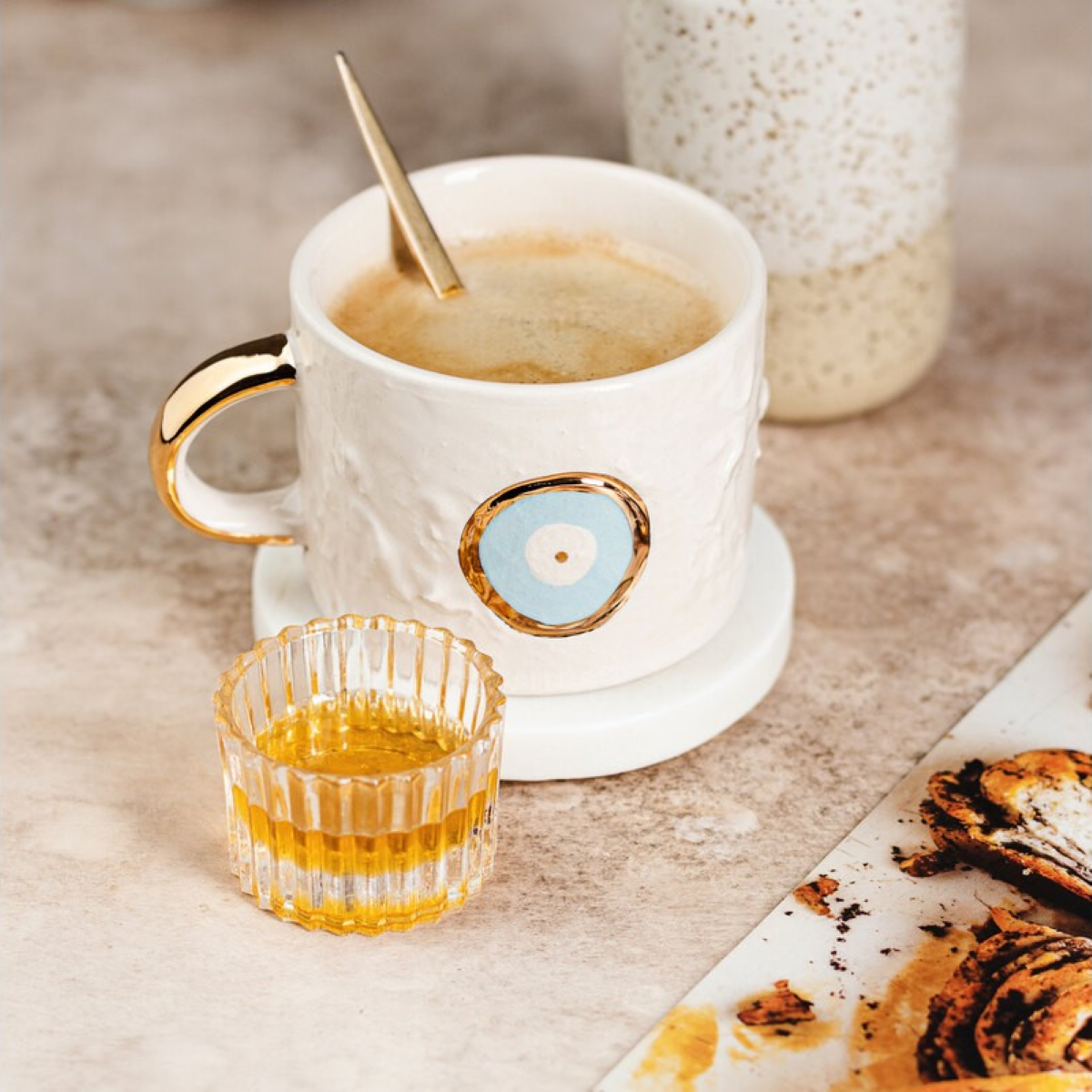 White mug with gold handle and decorative element, accompanied by a small glass of orange liquid on a beige surface.