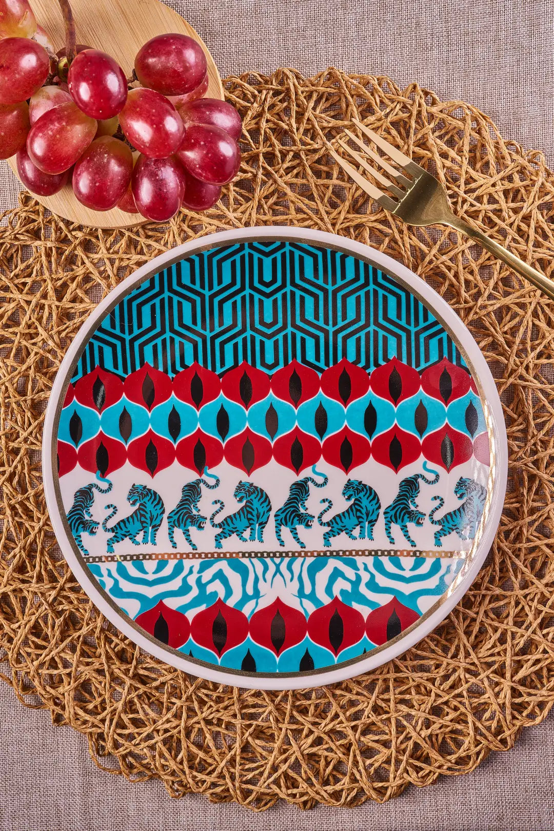 Gatto Wild porcelain salad plate with blue tigers and red geometric bands on a woven charger with grapes and a gold fork