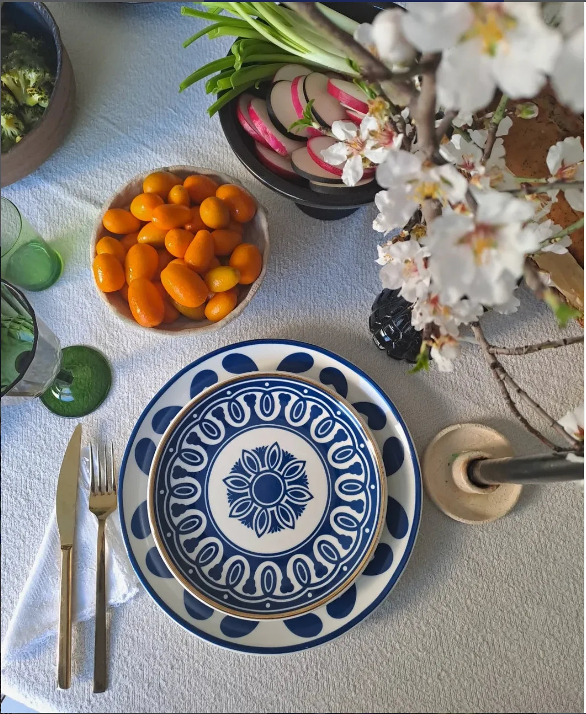 A styled tabletop setting featuring a blue and white ceramic plate with a floral medallion pattern, accompanied by gold-toned flatware, a bowl of orange kumquats, sliced radishes, green onions, and a white blossoming branch on a light blue tablecloth.