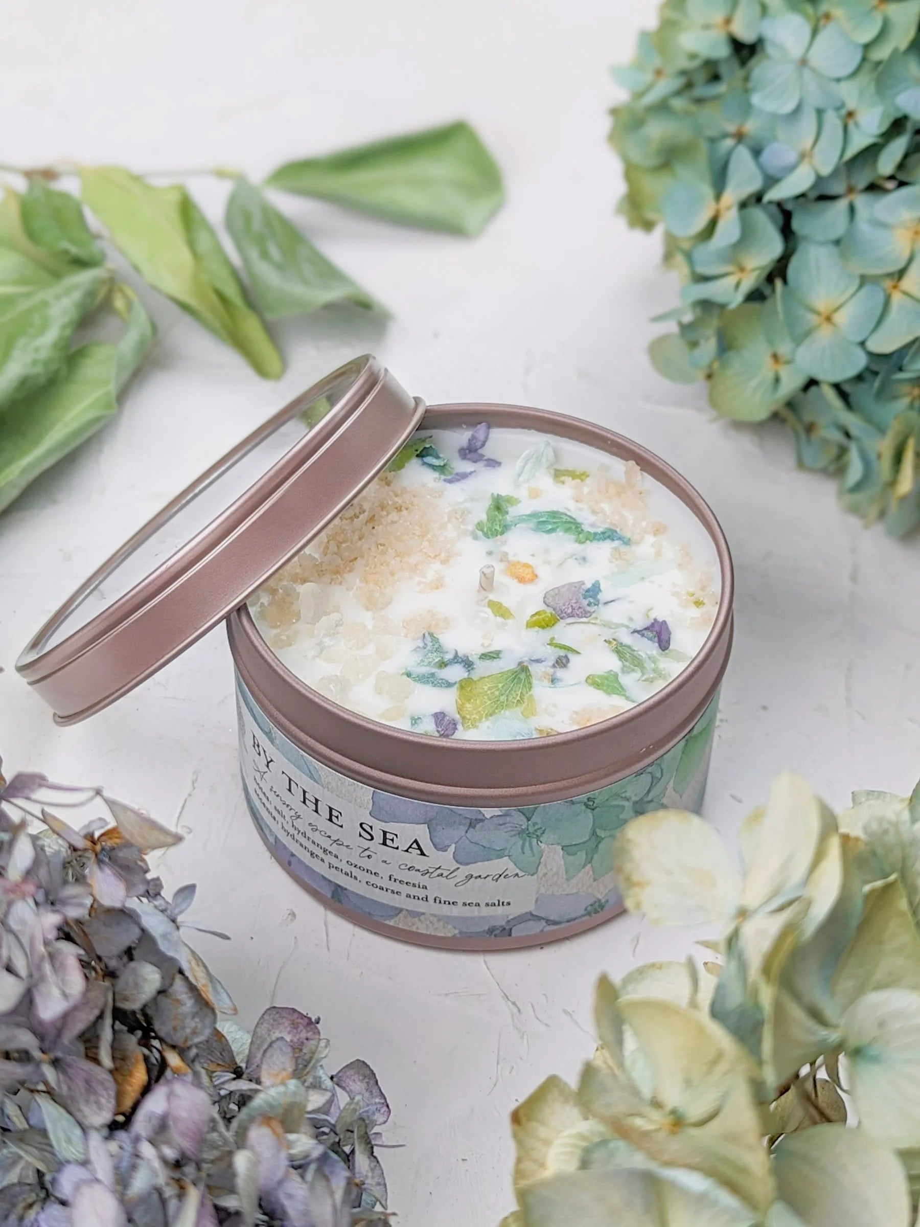 Candle in a tin with floral design on a white surface surrounded by flowers