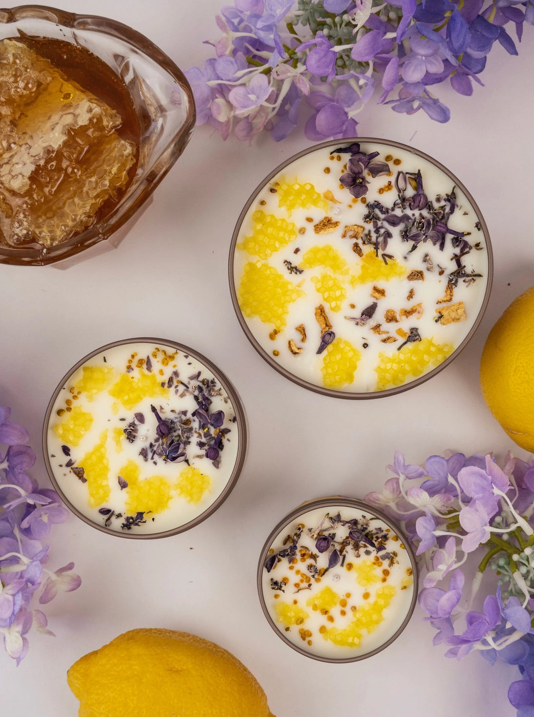 Three small bowls of candle with lemon and purple flowers, surrounded by lemons and flowers on a white background.