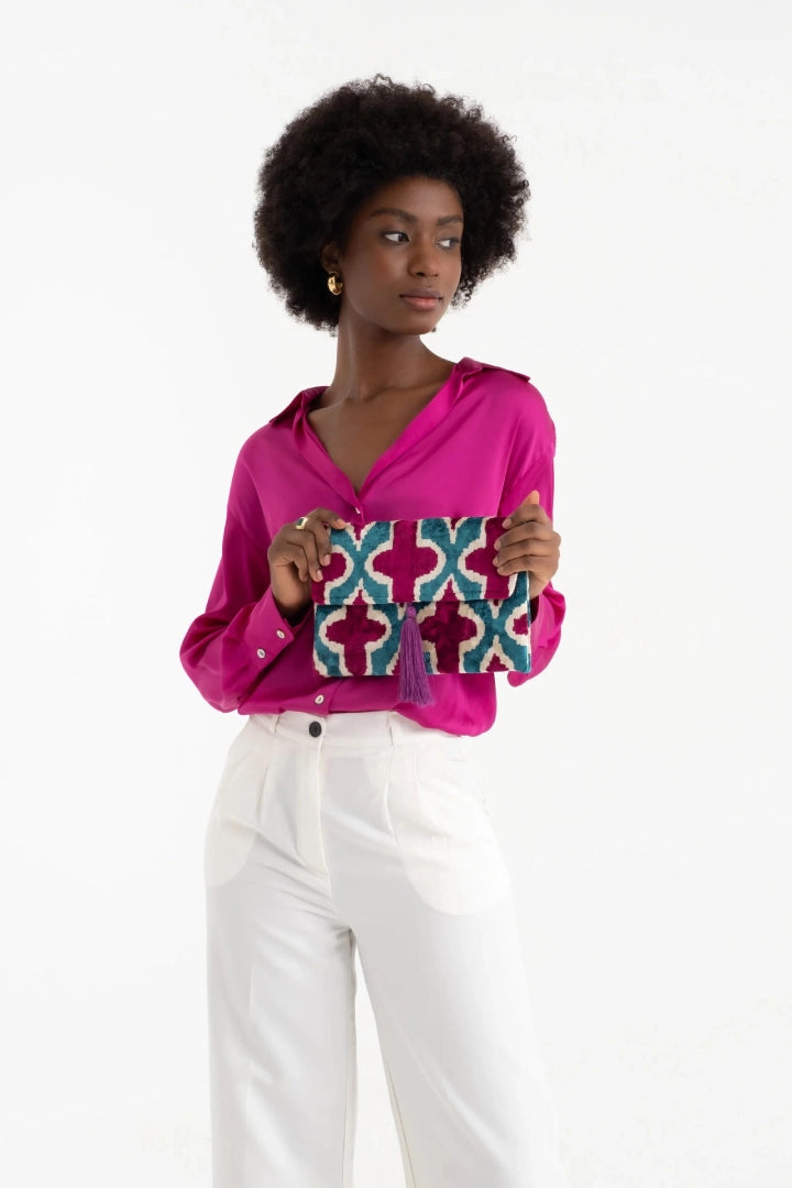 Model in a fuchsia shirt and white trousers holding the teal and magenta clutch